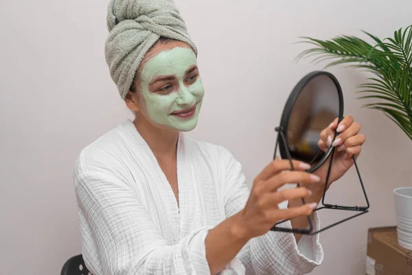 Cheerful woman with green clay mask on face looking in table mirror smiling lady in gown cleansing facial skin with homemade cosmetic product on pampering day