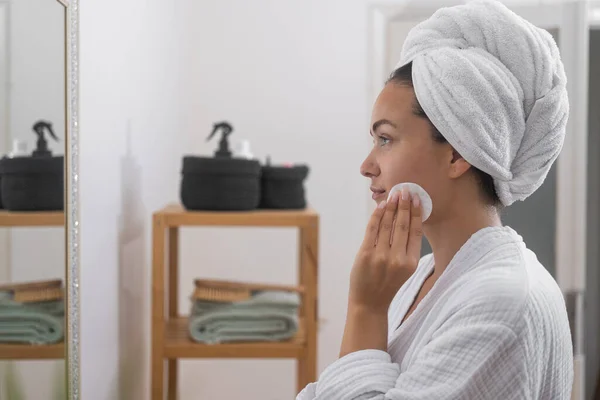 Woman cleaning facial skin with cotton disk near mirror in bathroom beautiful lady with towel turban taking care of skin with dermatological product at dressing table at home