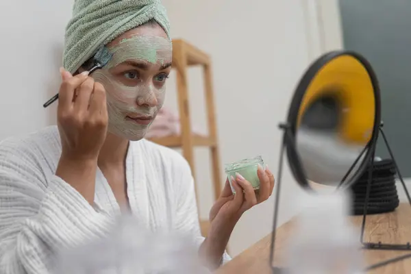 Woman applying homemade clay mask on face with brush sitting at table mirror lady in bathrobe using craft organic cosmetics to cleanse facial skin at home