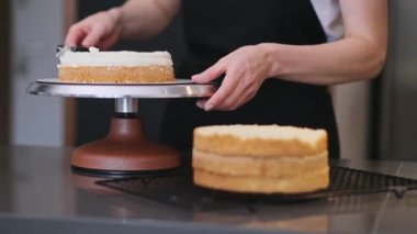 Taze pasta katmanlarına krema sürerek mutfak masasında oturan önlüklü kadın, evdeki etkinlikler için organik glütensiz hamur işi yapıyor.