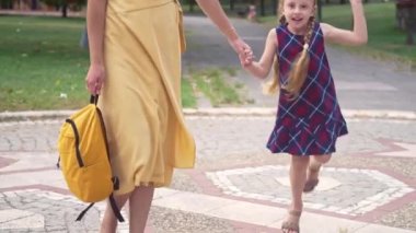 Aktif küçük kız annesiyle yeşil parktaki yeşil elbiseli kadını sırt çantasıyla geziyor ve neşeli kızını okula götürüyor.