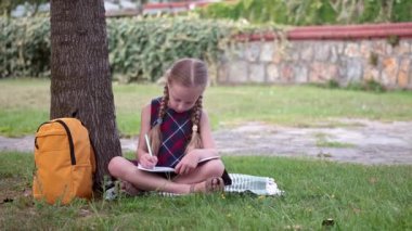 Saçı örgülü küçük bir kız okul bahçesindeki ağacın altında oturup ödev yapar. Okul öğrencisi, okul bahçesinde ders kitabı yazarak görevleri çözer.