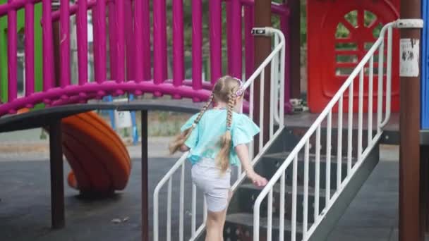 かなり小さな女の子は2階に上がり 子供の遊び場の幸せな子供の迷路でスライドし 公共の都市公園の子供の遊びのスライドがスライドを楽しんでいます — ストック動画