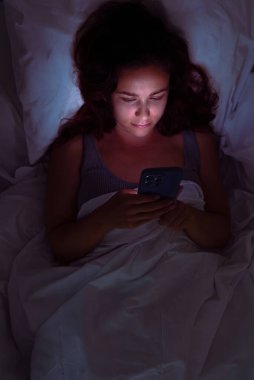 Karanlıkta saklanan bir kadın, yatakta telefonuna bakıyor, uykusuzluk ve sosyal medya saplantısına bir göz atıyor.. 