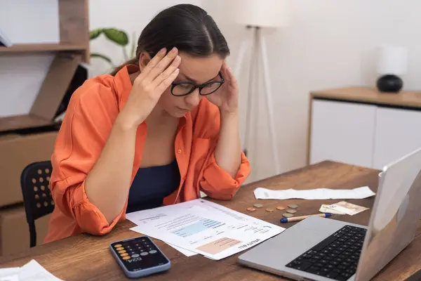 Stressed woman in glasses looks at paper bills feeling confused of high payment rates lady has problems while summarizing utilities sitting at table in office