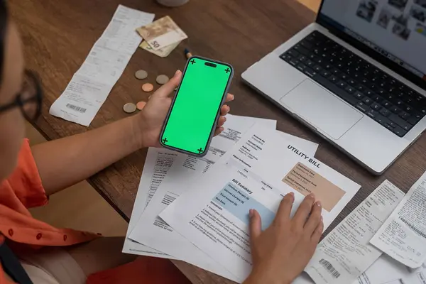 Digital Finance Hub: A womans hand grips a mobile phone with a chroma key screen on a wooden desk, surrounded by utility bills, money, and an online payment app.