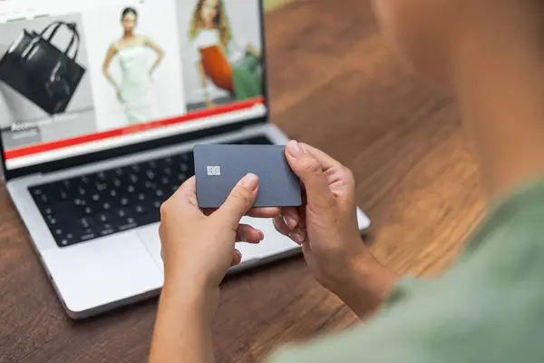 Female hands grip a credit card near a laptop featuring an online fashion stores website, symbolizing convenient online clothing shopping.