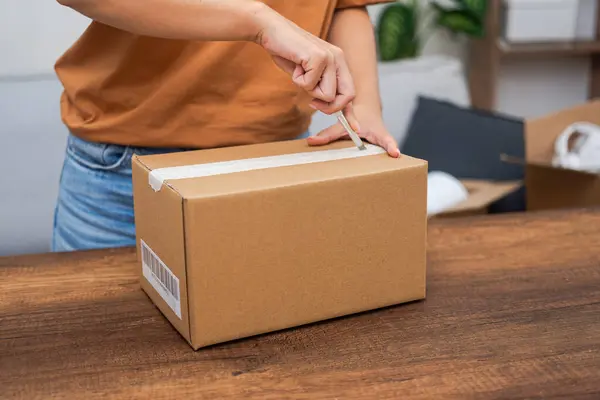 Housewife opens cardboard parcel with ordered goods with stationery knife on wooden table unpacking box on blurred background of open box and plant in pot in premise