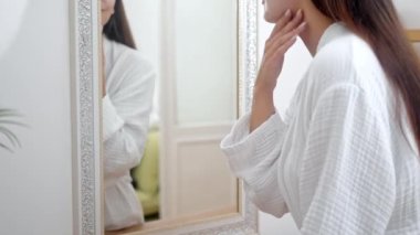 Bir kadın, rahat bornozuyla, evde güzel bir cilt bakımı yapıyor, banyo aynasında kendini inceliyor.. 