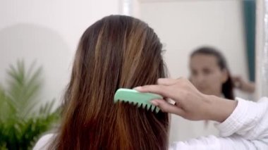 Ayna Mane: Genç bir kadın, aynanın önünde uzun ve sağlıklı saçlarını nazikçe tarıyor ve evde saç bakımını yapıyor.. 