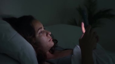 Geceleri kadın yatakta uzanıyor, telefon ekranı tarafından tüketiliyor, uykusuzluk ve akıllı telefon bağımlılığı tasvir ediyor.. 