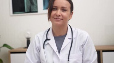 Ofisinde, gülümseyen bir kadın doktor, çevrimiçi bir randevu sırasında hastaları sıcak bir şekilde selamlayarak kameraya hitap eder.