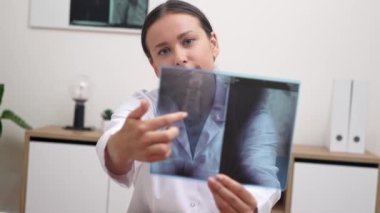  Kadın doktor bir teletıp muayenesi yapıyor, hastaya internetteki patolojiyi açıklarken bir röntgen filmini gösteriyor, kameraya bakıyor.. 