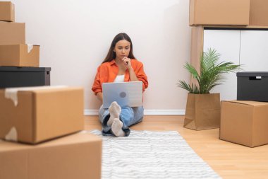 Elinde bir dizüstü bilgisayarla, genç bir kadın yeni bir daireye taşınmanın zorluklarını yönetiyor, paketlenmemiş kutularla çevrili.