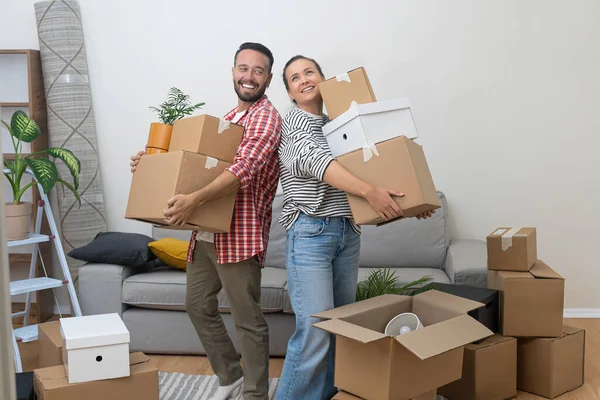 Relocation Challenges: A young couple, homeowners, struggles to hold a pile of cardboard moving boxes as they enter their new living room, determined to make it their own.