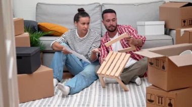 Mutlu genç çift, yeni apartmandaki kutularla çevrili modern ahşap mobilya montajı ve yeni ev dekorasyonu ile uğraşıyorlar.