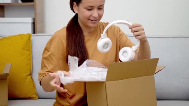彼女のソファーの幸せな女性は パッケージを開き 新しいヘッドフォンを発見し 彼女のオンライン購入に満足を表明 — ストック動画