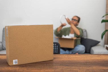 Bir masada, paket kutuları beklerken, arka planda, bir kadın gemicilik ve pazar alışverişini sembolize ederek online alışveriş paketini açıyor.