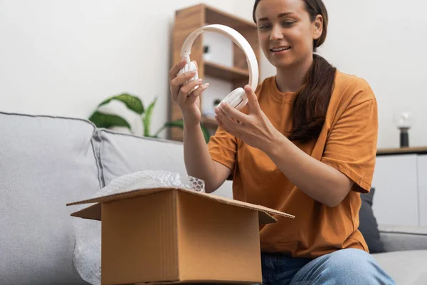 With joy, a woman unpacks her home-delivered package, revealing sleek modern headphones from an online store.