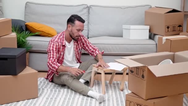 若い男が移動カートンボックスの中でDiyの棚を組み立て 移転後に新しい家で彼の資源を紹介する — ストック動画