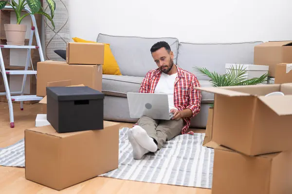 New Beginnings: Man sits amid boxes, laptop in hand, shopping online to decorate his fresh apartment, embracing the essence of moving in.
