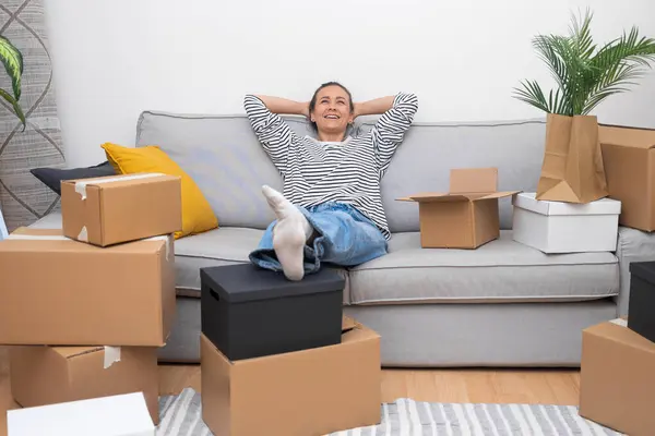 Embracing a joyful relocation, a content woman leans on her sofa, enveloped by unpacked boxes, signifying the real estate journey to her new, happy home
