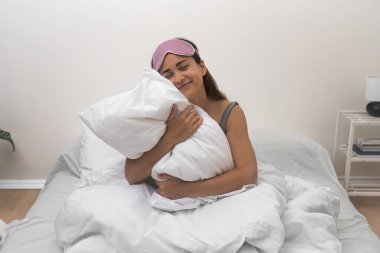 Çekici bir genç bayan, uyku maskesi takıyor, gülümsüyor, yatak odasında tüylü beyaz bir yastığı kucaklıyor, sabah mutluluğu yayıyor.. 