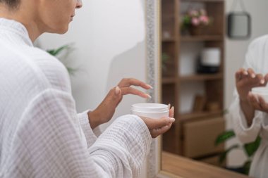 Beyaz bornozlu güzel bir kadın elinde bir kavanoz nemlendirici krem tutuyor.. 