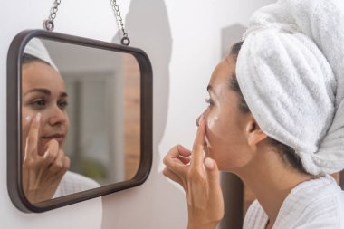 Aynadaki yansımasında, bir kadın cilt bakımını mükemmelleştirir, nemlendiriciyi uygular, günlük özbakımını üstlenir.. 