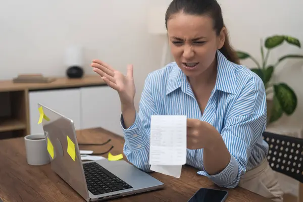 Angry woman looks at document with bank accounts frustrated by big utility bill lady waves hand aggressively sitting at workplace in office of apartment