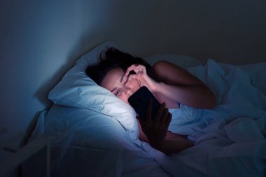 Yorgun bir kadın, yarı karanlık bir yatak odasında uykusundan önce telefon ekranında parıldıyor.