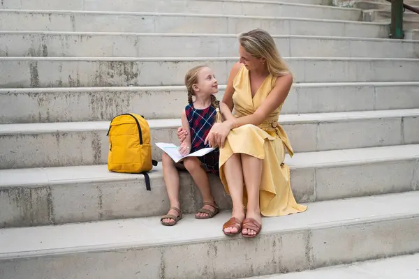 Cheerful mother and daughter do homework together sitting on high stairs in park. couple communicates and looks at each other with love hugging gently