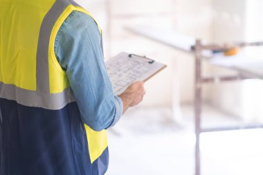 Yelek müteahhiti, inşaat güvenlik ekipmanlarının yanında duran profesyonel üniformalı pano işçisinin yenileme projesini kontrol ediyor.