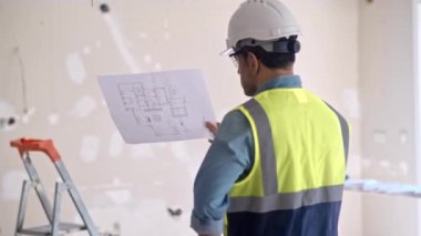 Çelik yelek giyen bir adam, yenileme işlemine başlamadan önce dairedeki eski püskü duvarın karşısında duran üniformalı bir uzman tarafından çizim kağıdını kontrol ediyor.