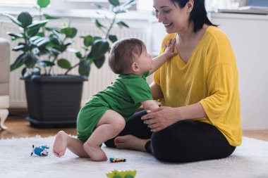 Sevimli erkek bebek annesine emekliyor dağınık oyuncakların yanında beyaz halıya oturuyor mutlu anne gülümsüyor küçük oğlunun evde çocuk odasında vakit geçirmesini seyrediyor.