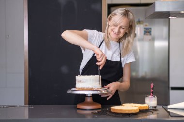 Tatmin olmuş pastacı gülümseyerek kek tabaklarını siyah önlüklü, organik krema sarışınla kaplarken mutfak masasında müşteriler için hamur işi pişirmenin keyfini çıkarıyor.