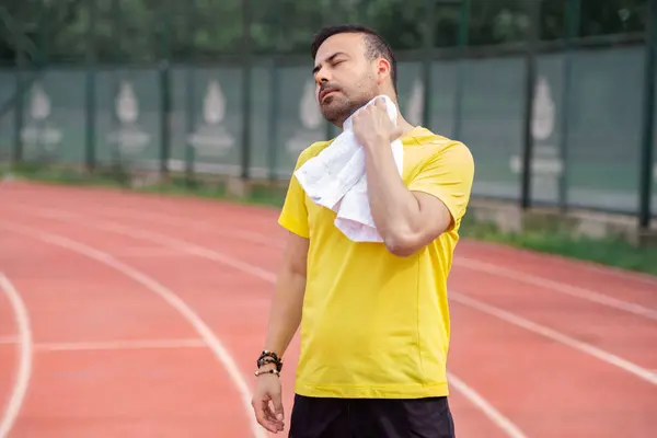 Exhausted sportsman wiping sweaty neck with soft towel standing on running track after jogging training at urban stadium athlete resting after workout on outdoor arena