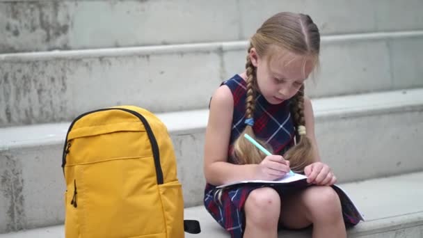 勤奋的小女孩坐在书架上 靠着书包坐在石阶上 可爱的小女孩背着书包在学校院子里做作业 — 图库视频影像