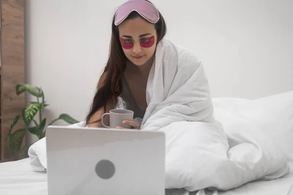 A lady with eye patches and coffee, nestled in bed, efficiently manages online work and remote studies using her laptop from home.