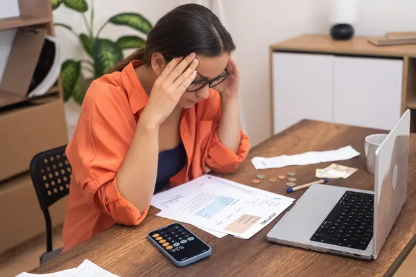 Financial Confusion: In glasses, a stressed woman at a table looks at paper bills, perplexed by the high payment rates.