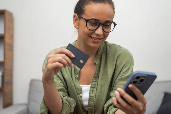 Positive woman makes online payment for purchases with mobile phone holding credit card in hand concept of ordering goods for home delivery and innovation technology