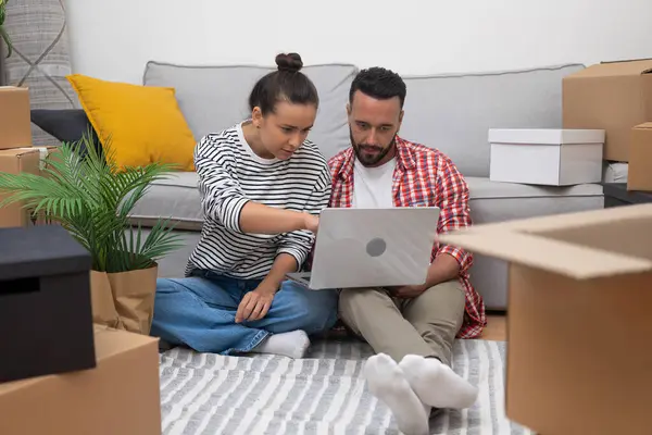 Couple sits on floor among unpacked boxes together choose wallpapers for new kitchen from web site on laptop and wife shows favorite wallpaper to husband