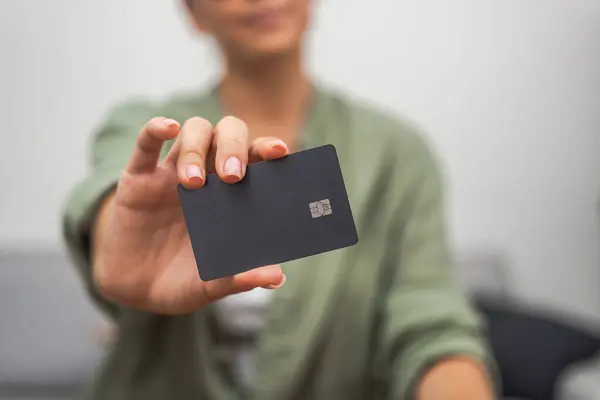 Lady shows dark credit card with chip for contactless payment sitting on sofa in accommodation concept of online payment in Internet store at home and technology