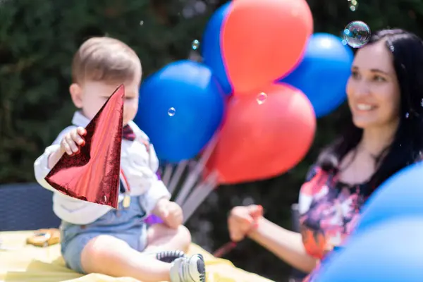Bubble-filled joy on the first birthday, baby with Happy Birthday hat outdoor celebration featuring red and blue balloons, bubble blowing