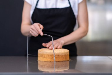 Siyah önlüklü kadın mutfak masasında el yapımı pasta yapıyor. Katmanları ayırmak için fırında pişirilmiş şekerlemeleri tel bıçakla kesiyor.
