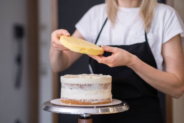 Glütensiz pasta katmanları bağlayan siyah önlüklü profesyonel pastacı. Beyaz kremalı yetenekli sarışın kadın müşteriler için el yapımı düğün pastası üzerinde çalışıyor.