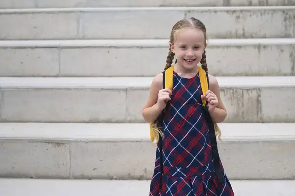In her school dress and yellow backpack, a happy blonde schoolgirl smiles with pure joy as she embarks on her journey to school