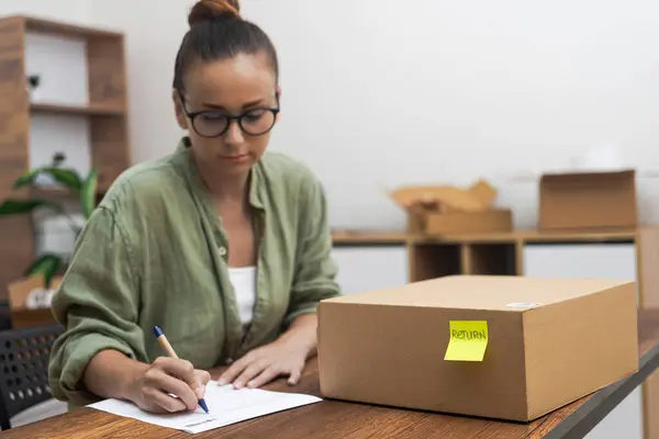 Serious manager in glasses writes application to send cardboard box with yellow sticker with goods back woman issues return to online store sitting at desk with parcel