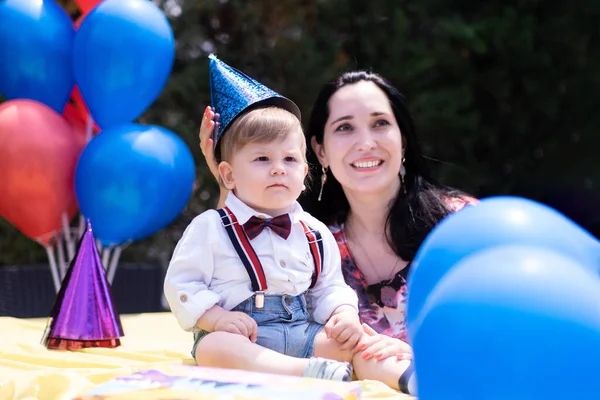 Mother son first year birthday, toddler style with suspenders, first birthday, balloons, party hats, and an assortment of birthday decorations.