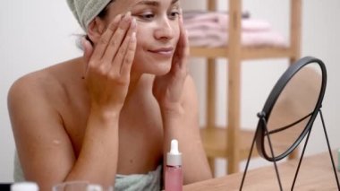 Sakin güzellik: Kafası havluya sarılı bir kadın, aynalı masada rahatlatıcı cilt bakımı için hyaluronik asit serumu kullanır.. 